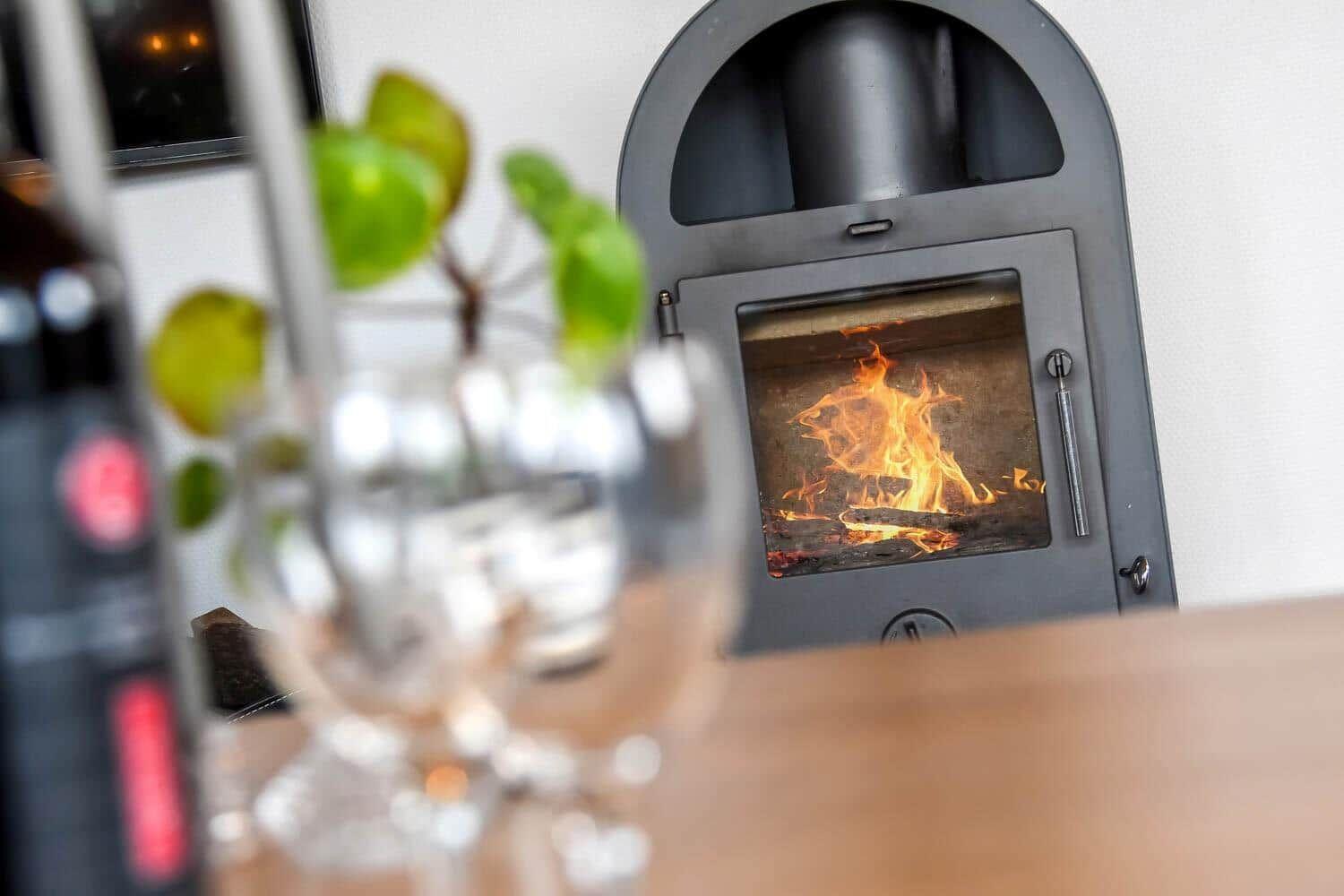 Ein Kaminofen mit brennendem Feuer, im Vordergrund unscharf ein Glas und eine Pflanze.