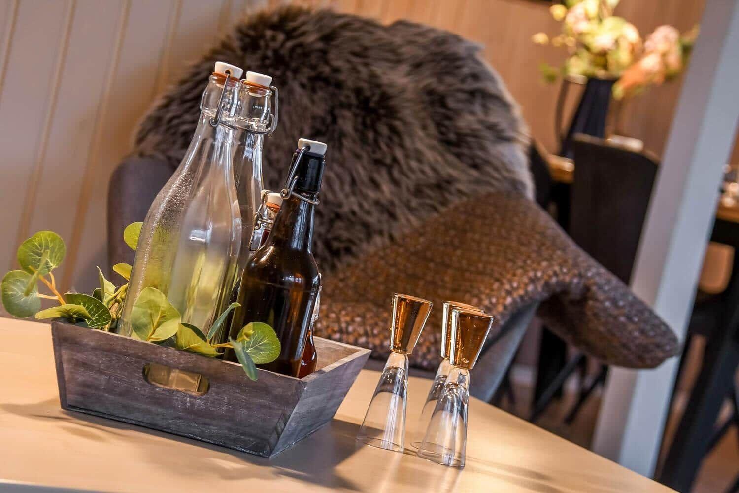 Holztablett mit Flaschen und Gläsern auf Tisch, Hintergrund mit Felldecke und Blumen.