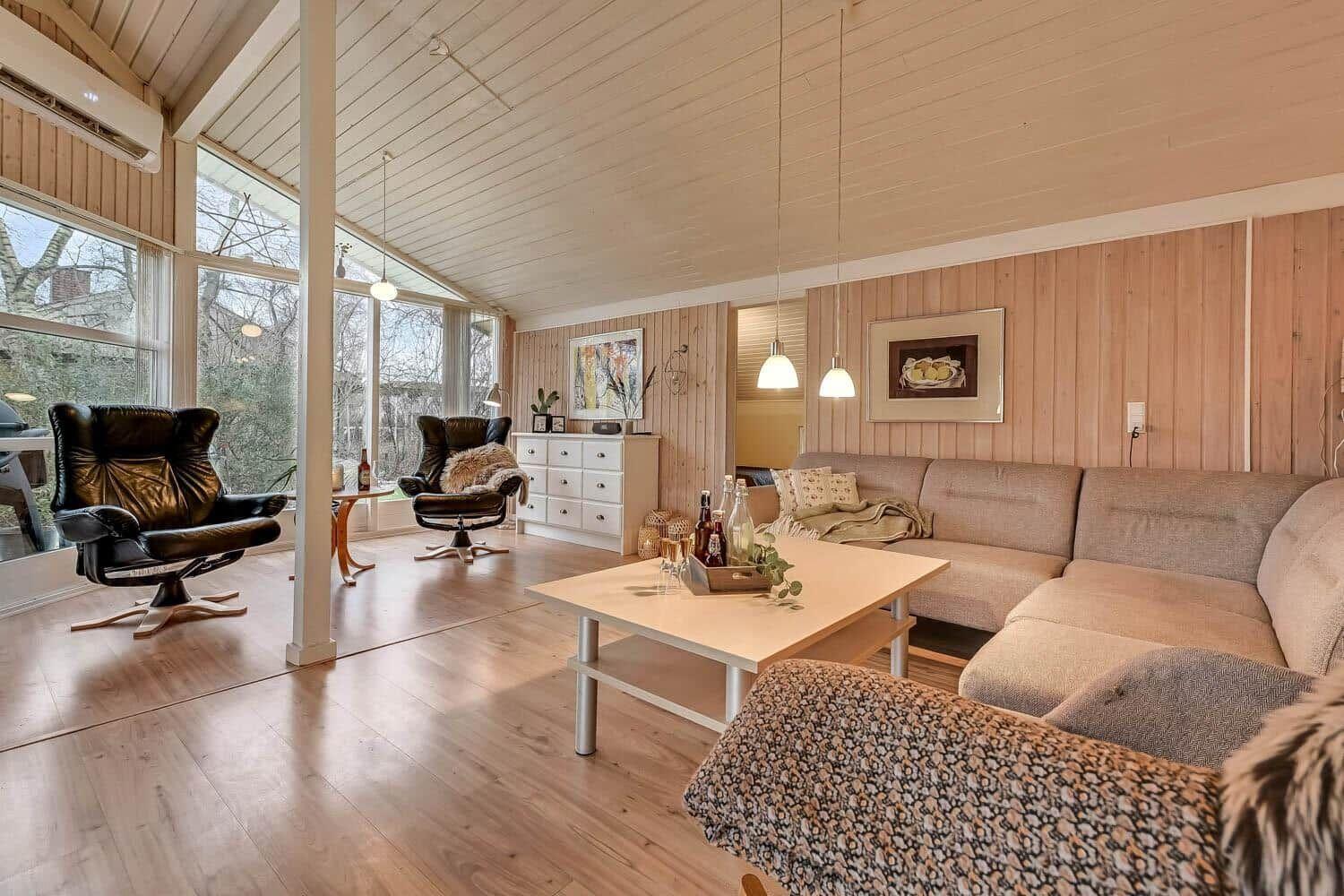 Wohnzimmer mit Sofa, zwei Sessel und großem Fenster. Holzdecke und -boden.