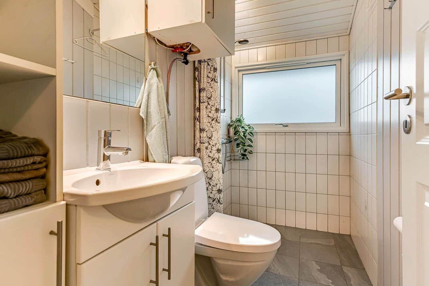 Badezimmer mit Waschbecken, Toilette und Dusche. Fenster und Pflanze sichtbar.
