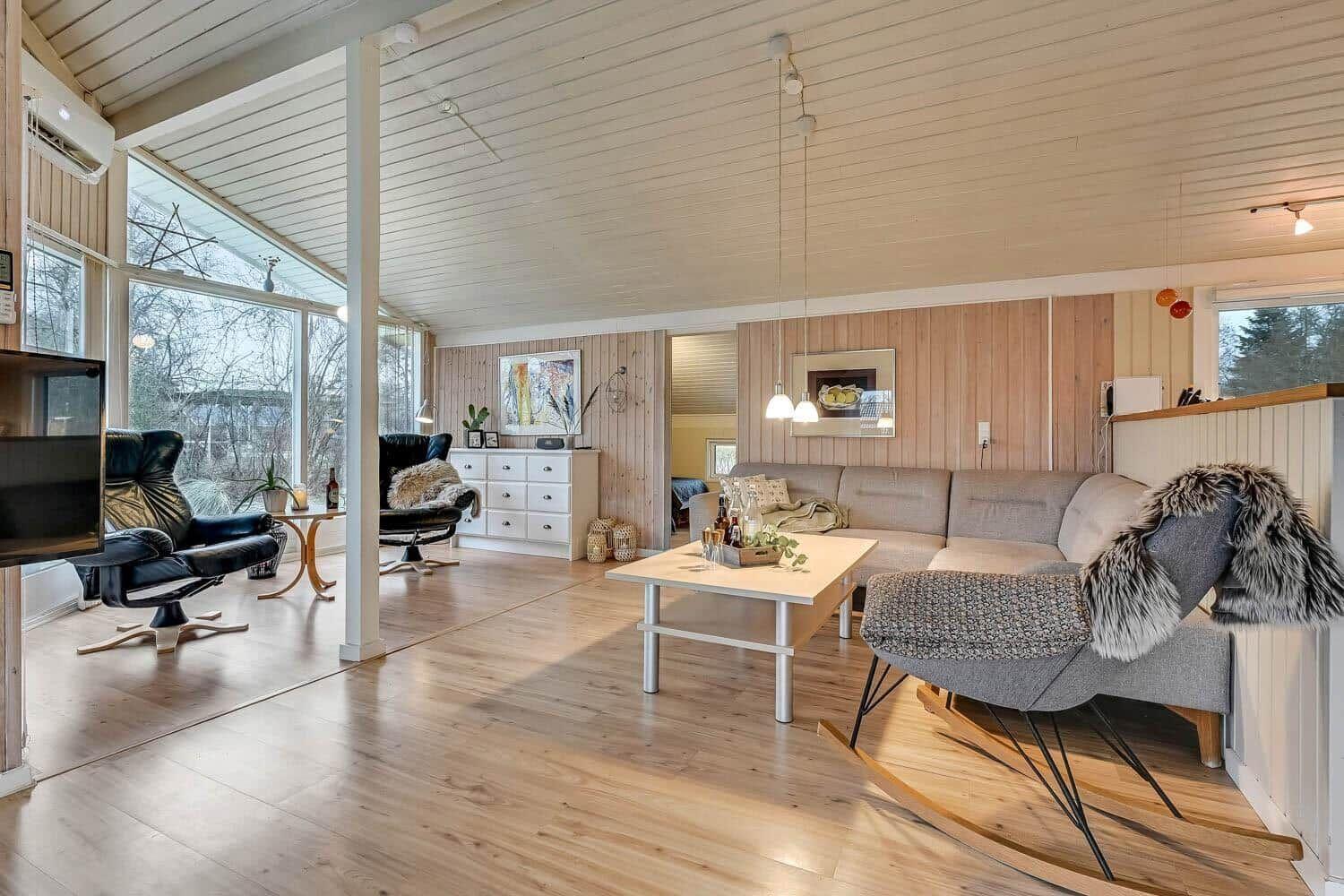 Wohnzimmer mit Sofa, Schaukelstuhl und Holzboden. Große Fenster und Holzwände.