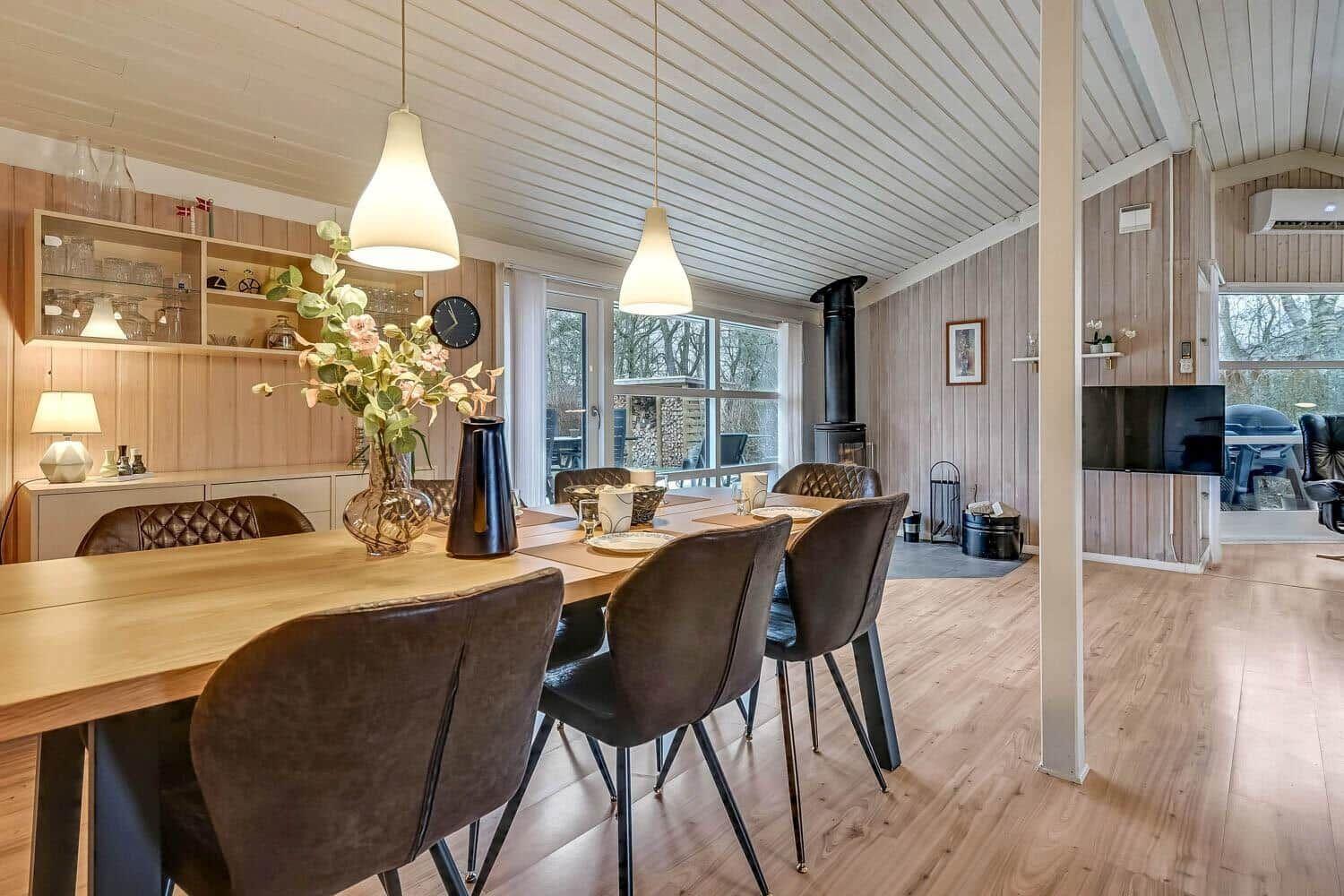 Esszimmer mit Holztisch, Stühlen und Fenstern. Hintergrund: Wohnbereich mit Kamin und Fernseher.