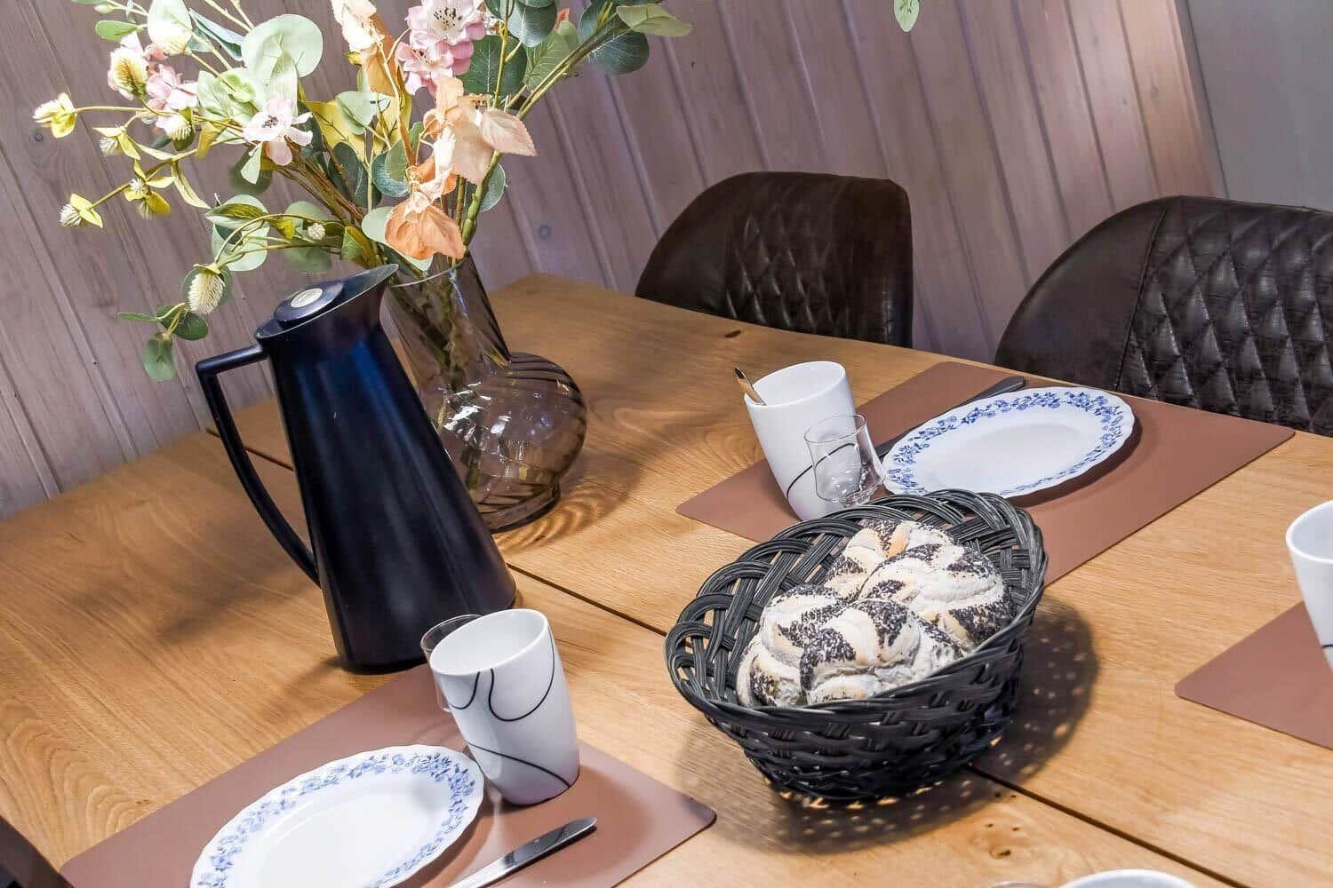 Holztisch mit Stühlen, Teller, Kaffeekanne und Brotkorb.