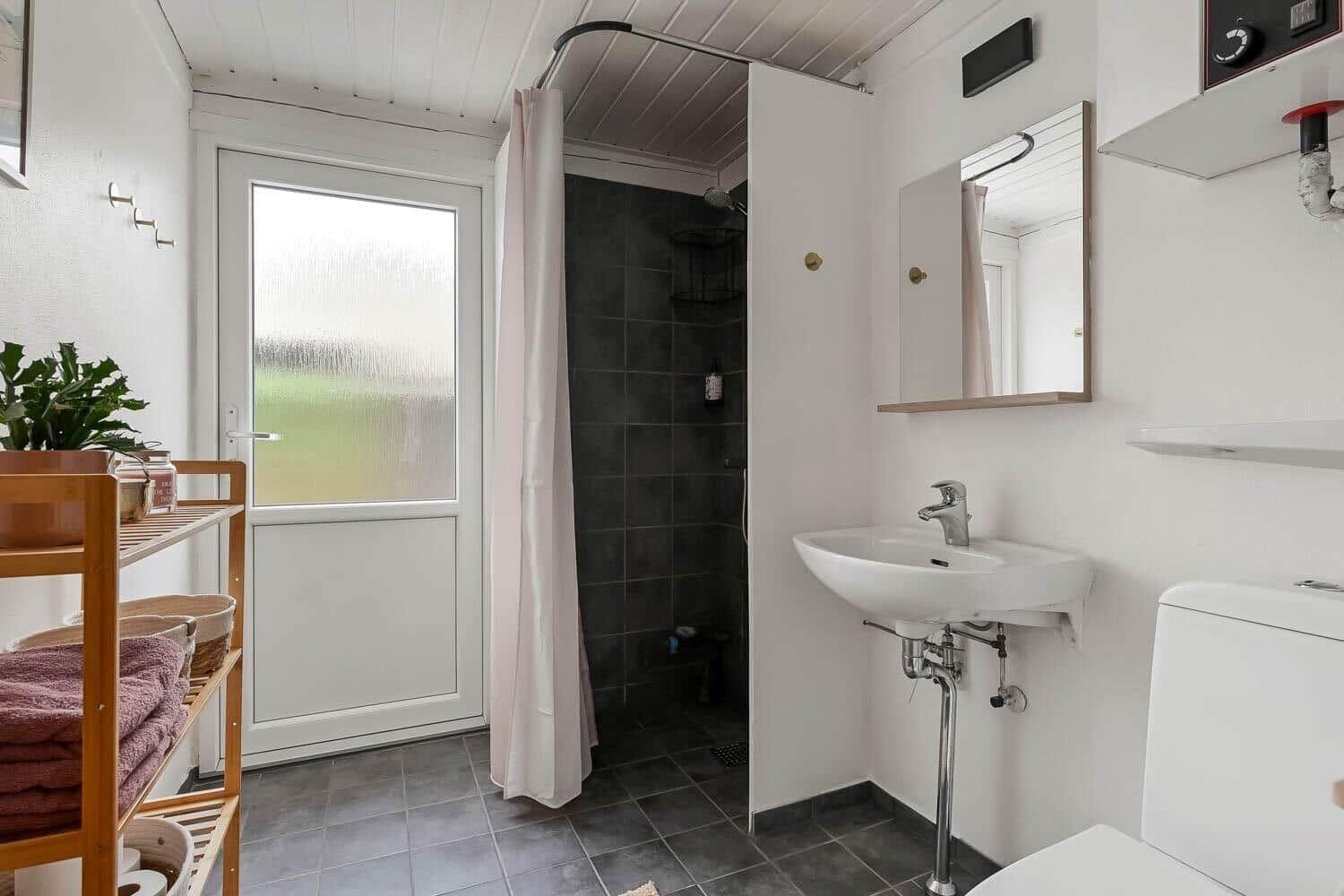 Badezimmer mit Dusche, Waschbecken und Toilette. Holzregal mit Handtüchern und Pflanze.