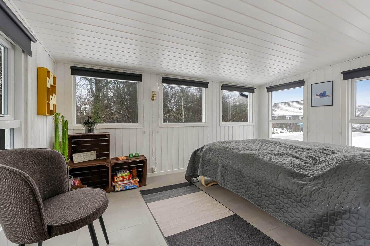 Schlafzimmer mit Bett, Stuhl und Fenstern. Wand mit Holzverkleidung und Einrichtungsgegenständen.