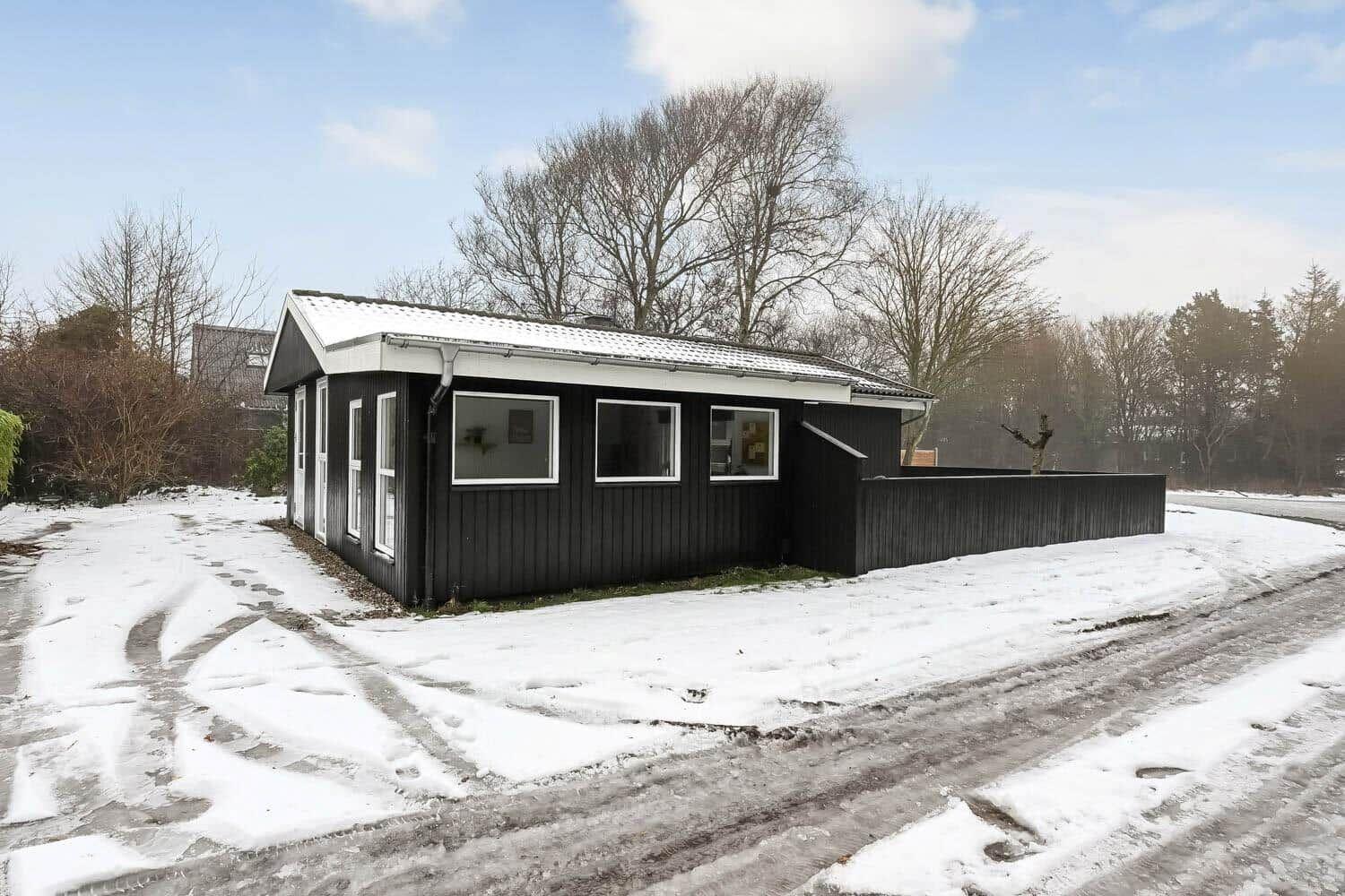 Schwarzes Holzhaus mit weißem Dach und Fenstern, umgeben von Schnee und Bäumen.