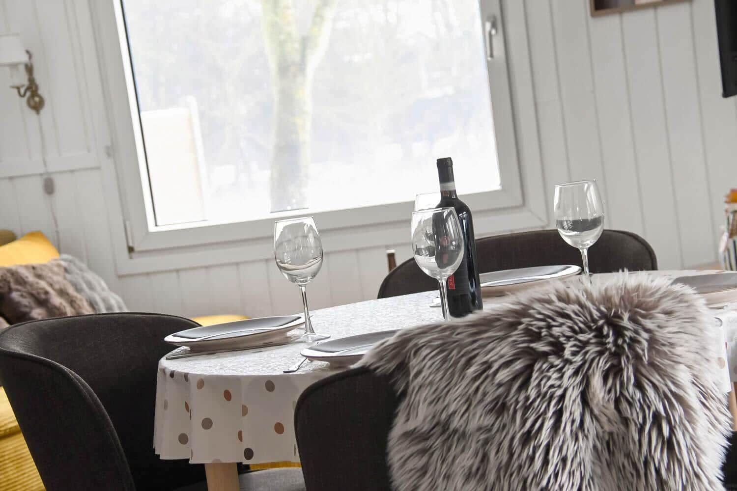Esstisch mit Tischdecke, Gläsern und Flasche vor Fenster.