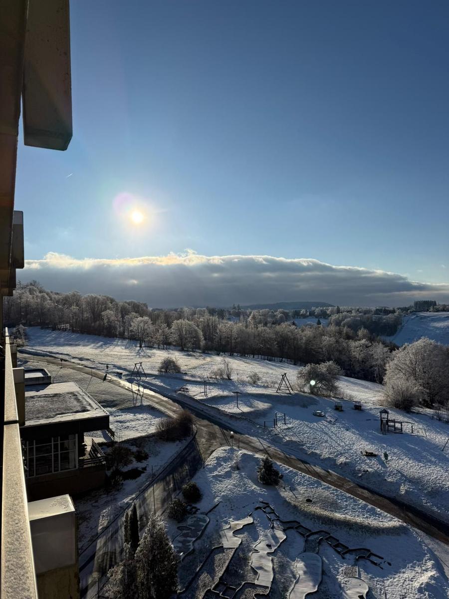 Schneebedeckter Blick auf einen Park mit Spielplatz und Bäumen unter blauem Himmel.