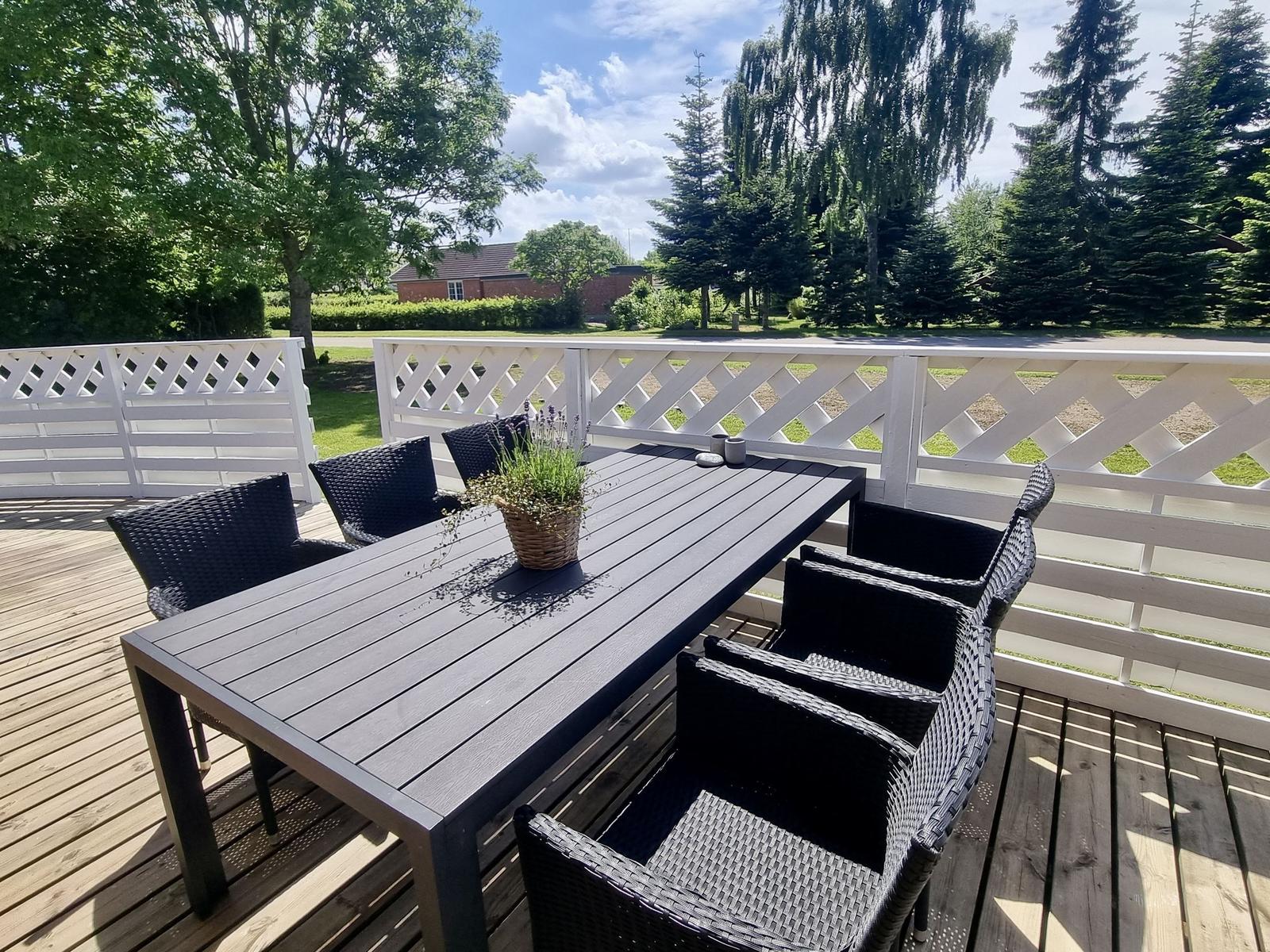 Terrasse mit Tisch und sechs Stühlen, weißer Zaun, Blick auf Grünflächen und Bäume.