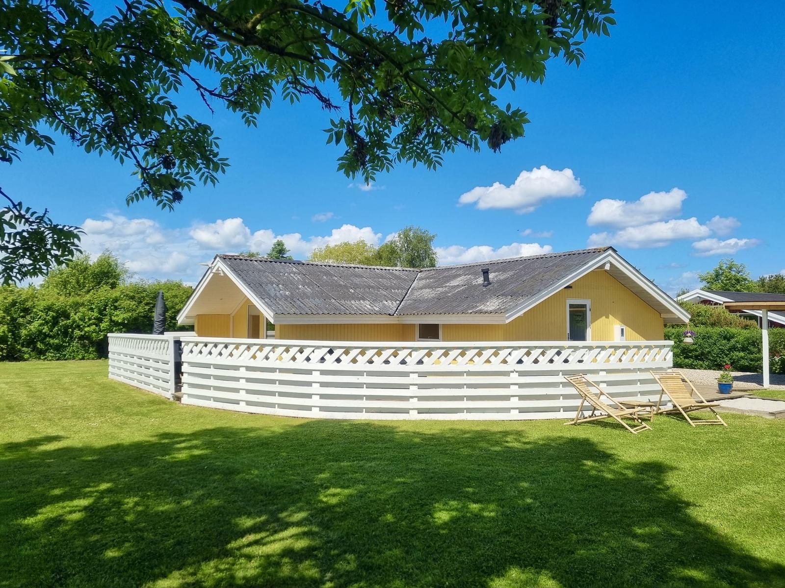 Ein gelbes Haus mit weißer Umzäunung und Gartenliegen unter blauem Himmel.
