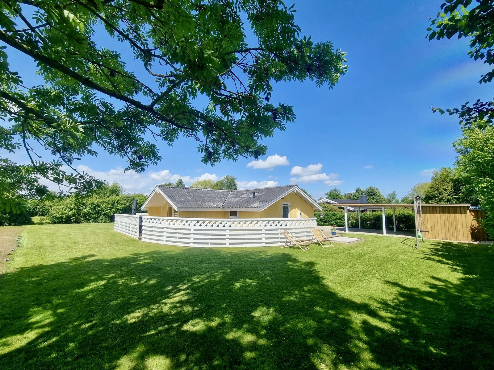 Ein gelbes Haus mit Garten, weißer Umzäunung und Liegestühlen unter Bäumen.