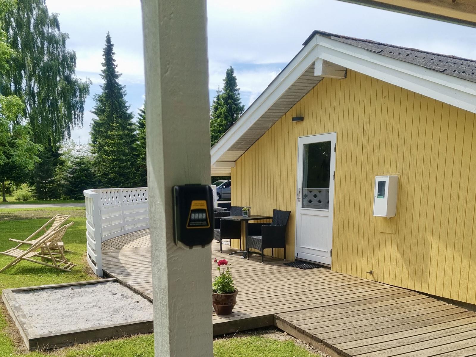 Haus mit Terrasse, Tisch, Stühlen und Sandkasten. Hintergrund: Gras, Bäume.