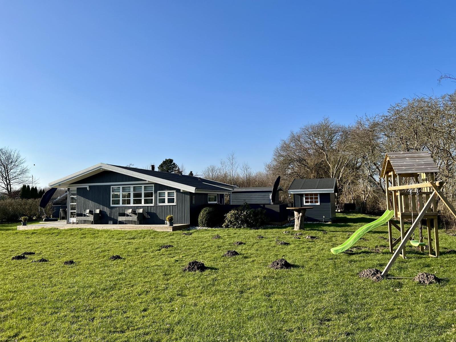 Ein graues Ferienhaus mit großem Garten und Kinderspielplatz unter blauem Himmel.