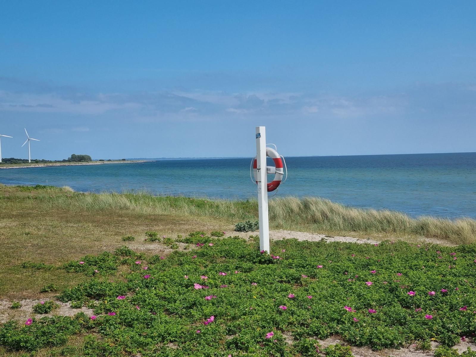 Rettungsring an Pfosten am Strand mit blauem Meer und Windkraftanlagen im Hintergrund.