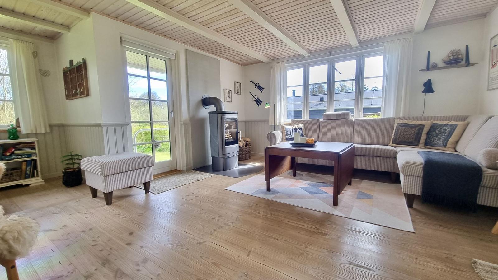 Wohnzimmer mit Sofa, Kaminofen und Holzboden. Große Fenster und Türen mit Blick auf Garten.