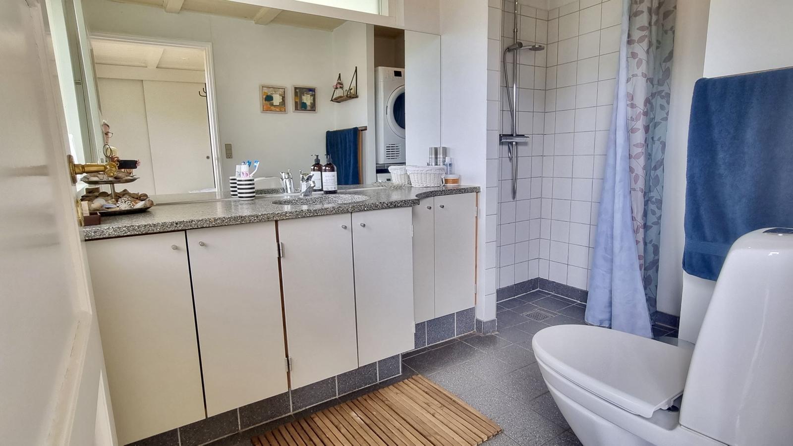 Badezimmer mit Dusche, Waschbecken und Toilette. Fliesenboden und Holzmatte.