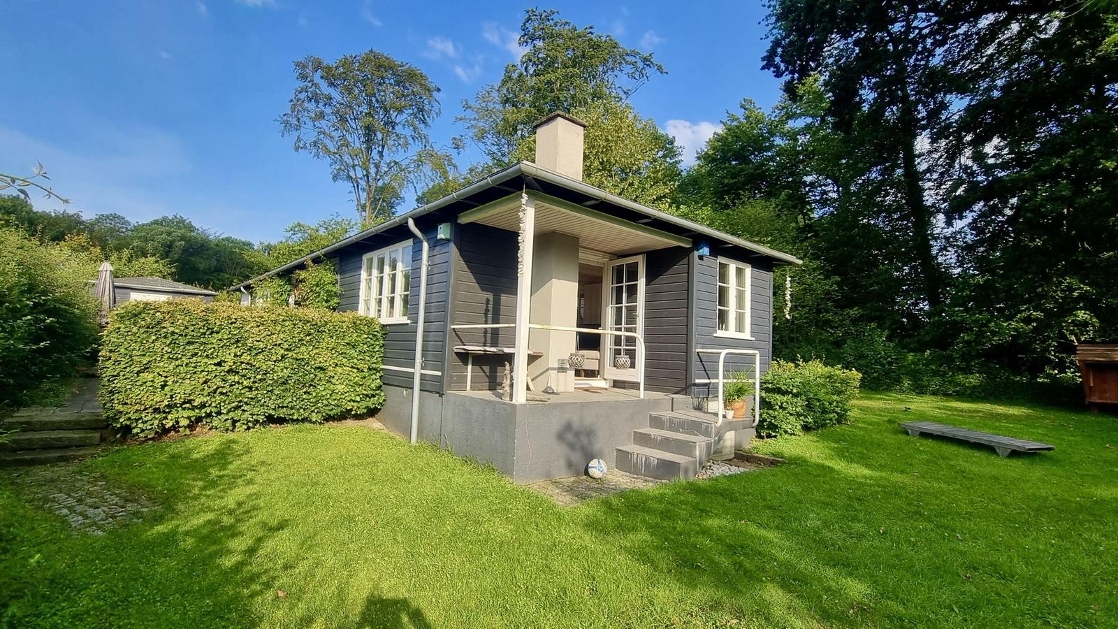 Ein graues Haus mit Veranda und Garten. Umgeben von Bäumen und Gras.