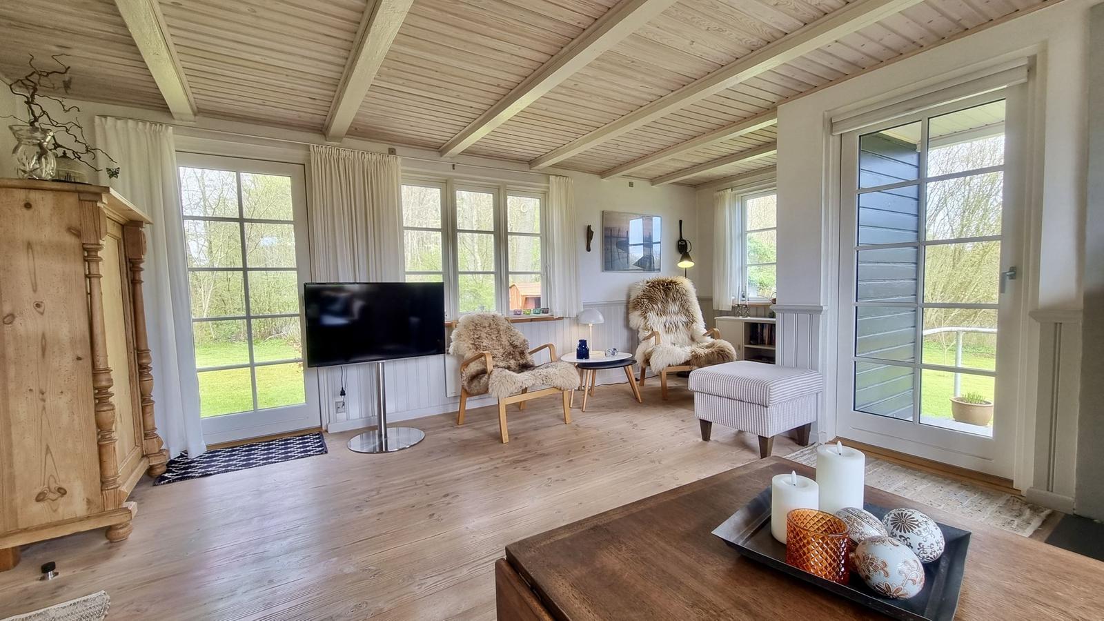 Wohnzimmer mit Holzboden, TV, zwei Sessel und großen Fenstern.