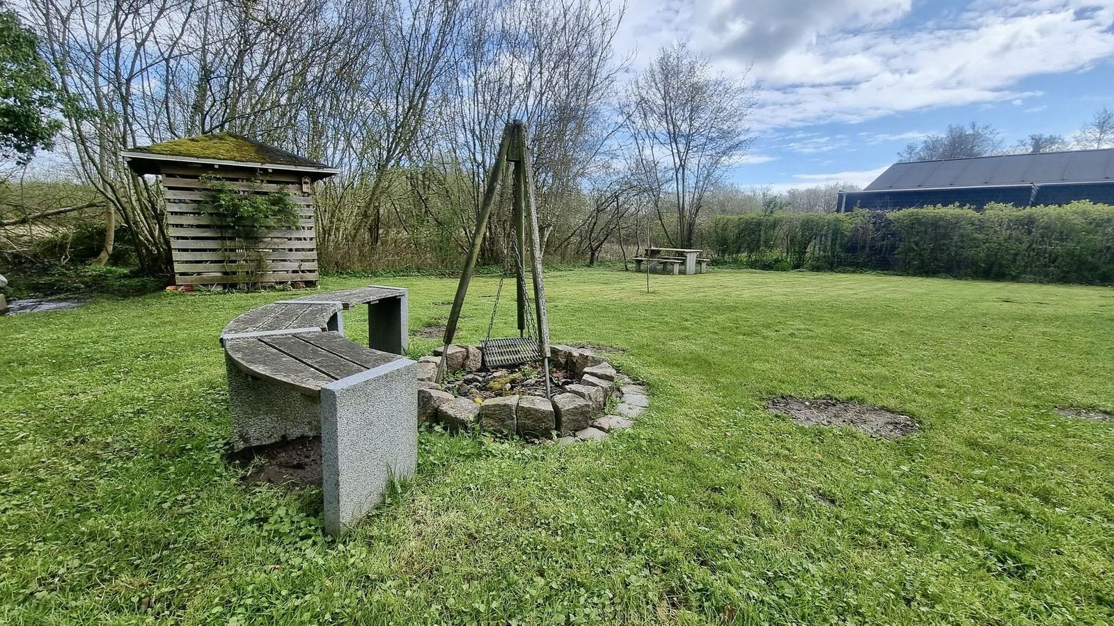 Grüner Garten mit Steinbank, Brunnen und Holzschuppen. Im Hintergrund steht ein Gebäude.