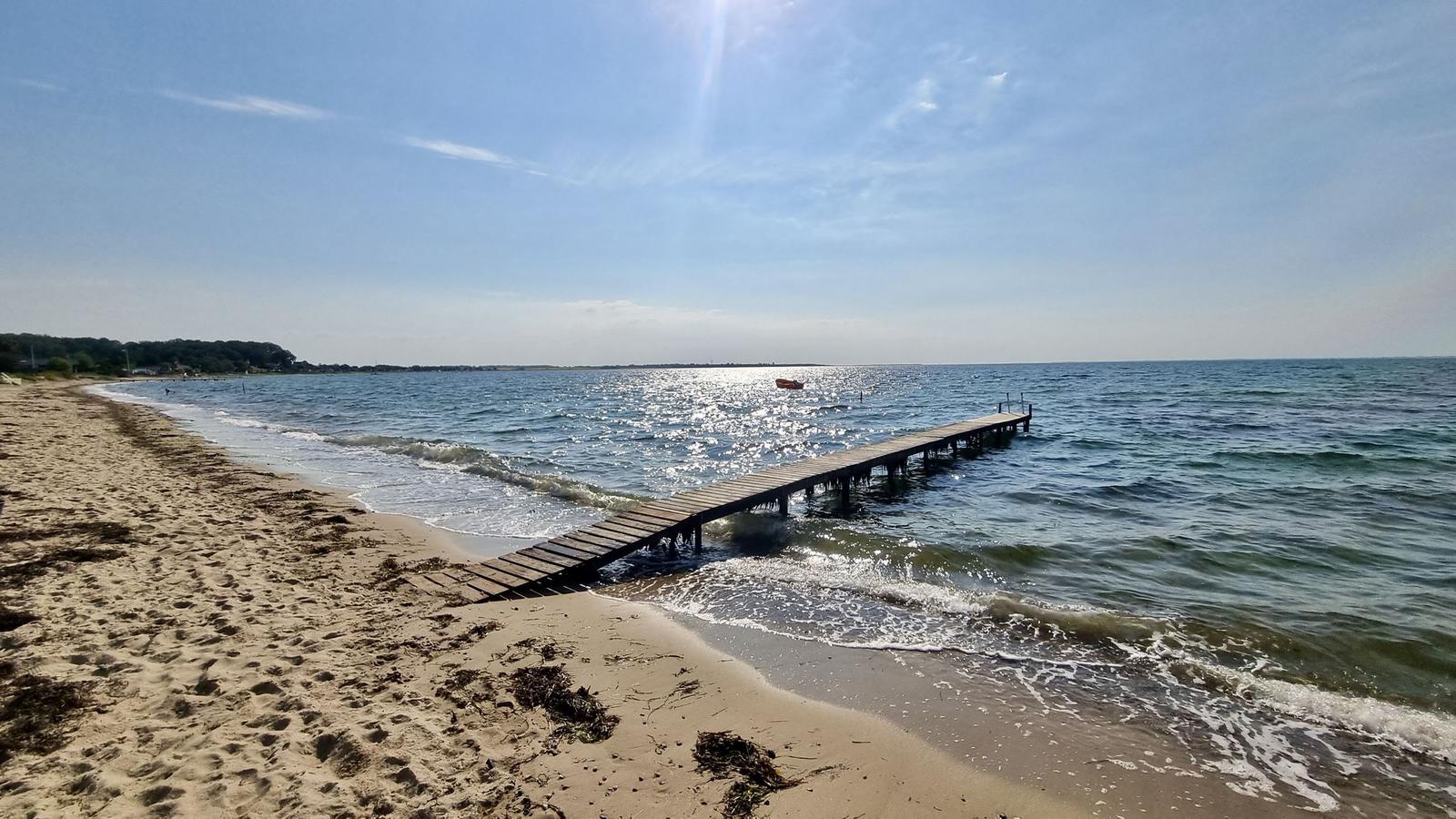 Holzsteg führt vom Strand ins Meer. Boot liegt im Wasser. Sonne scheint.
