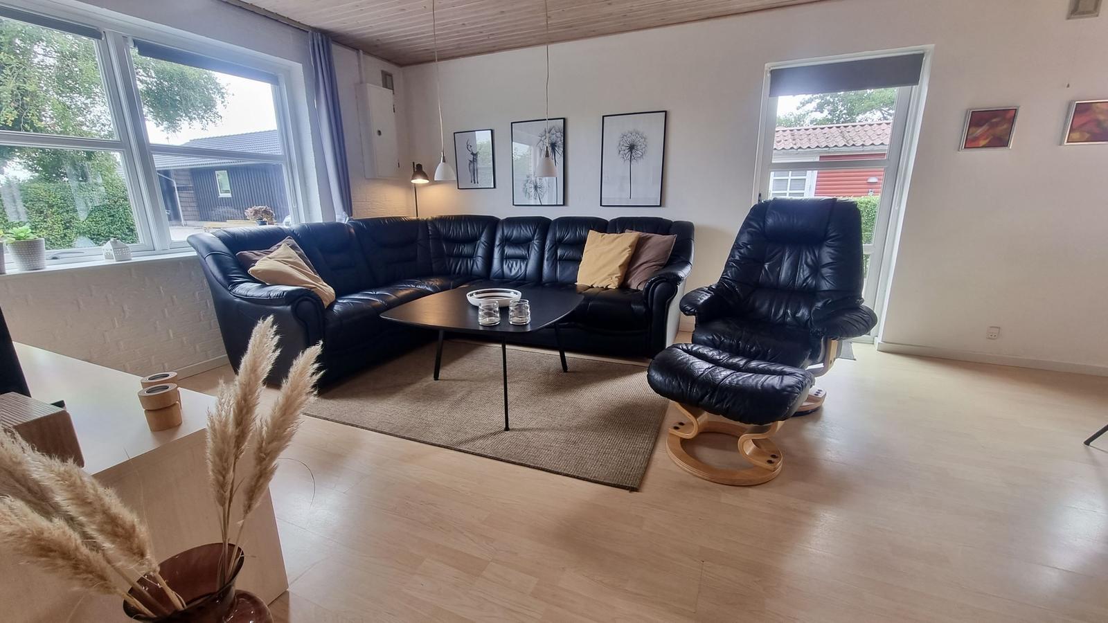 Wohnzimmer mit schwarzer Ledercouch, Sessel und Holzboden. Fenster und Tür bieten Blick nach draußen.