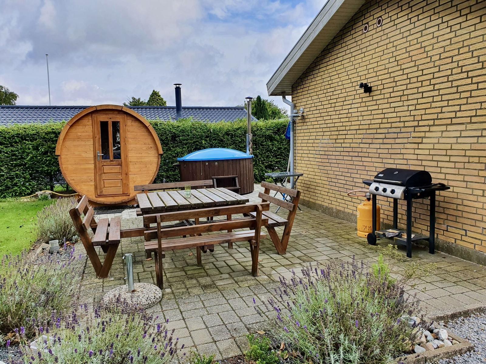 Terrasse mit Holztisch, Grill und runden Holzhaus. Lavendelbeete und Steinplatten.