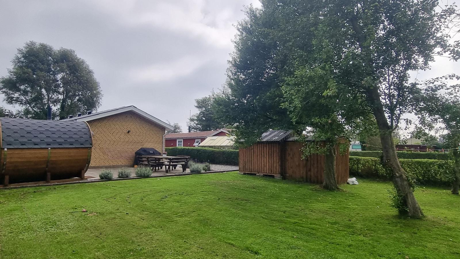Grüner Garten mit Holzsauna, Grill und Picknicktisch. Hintergrund: Haus und Bäume.