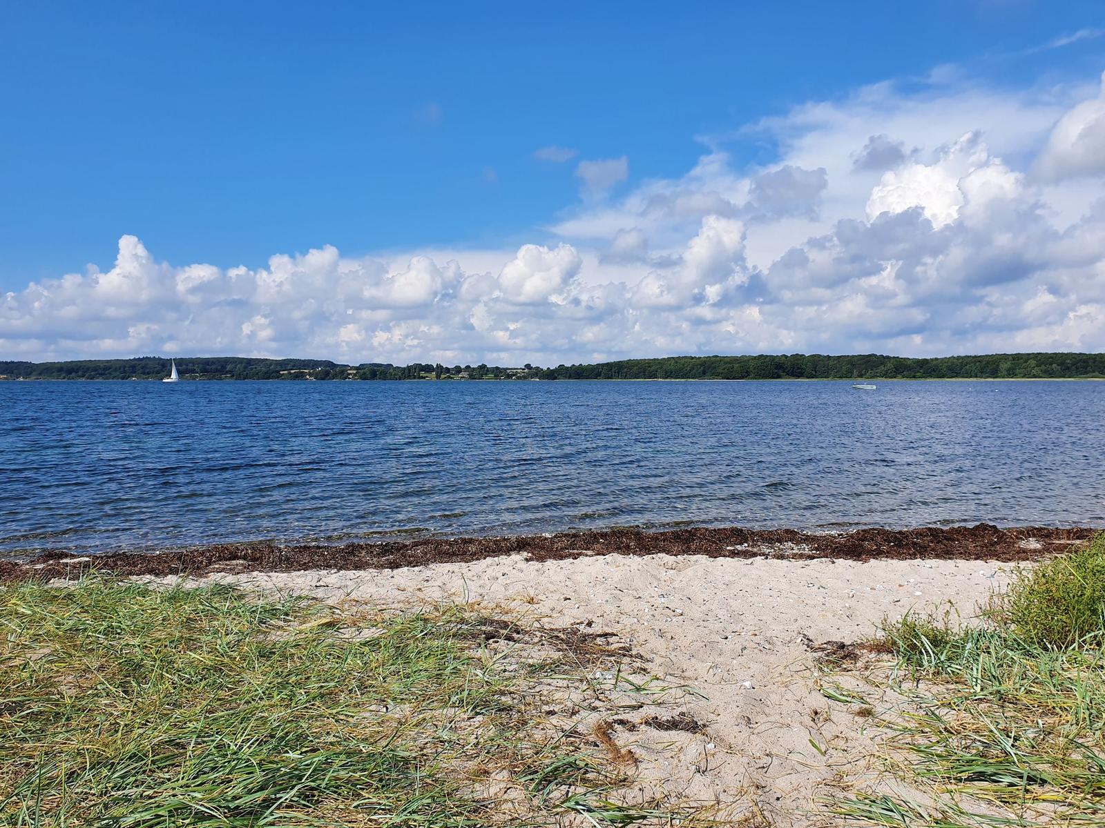 Strand mit Sand und Gras, See mit Segelboot und Boot im Hintergrund unter blauem Himmel mit Wolken.