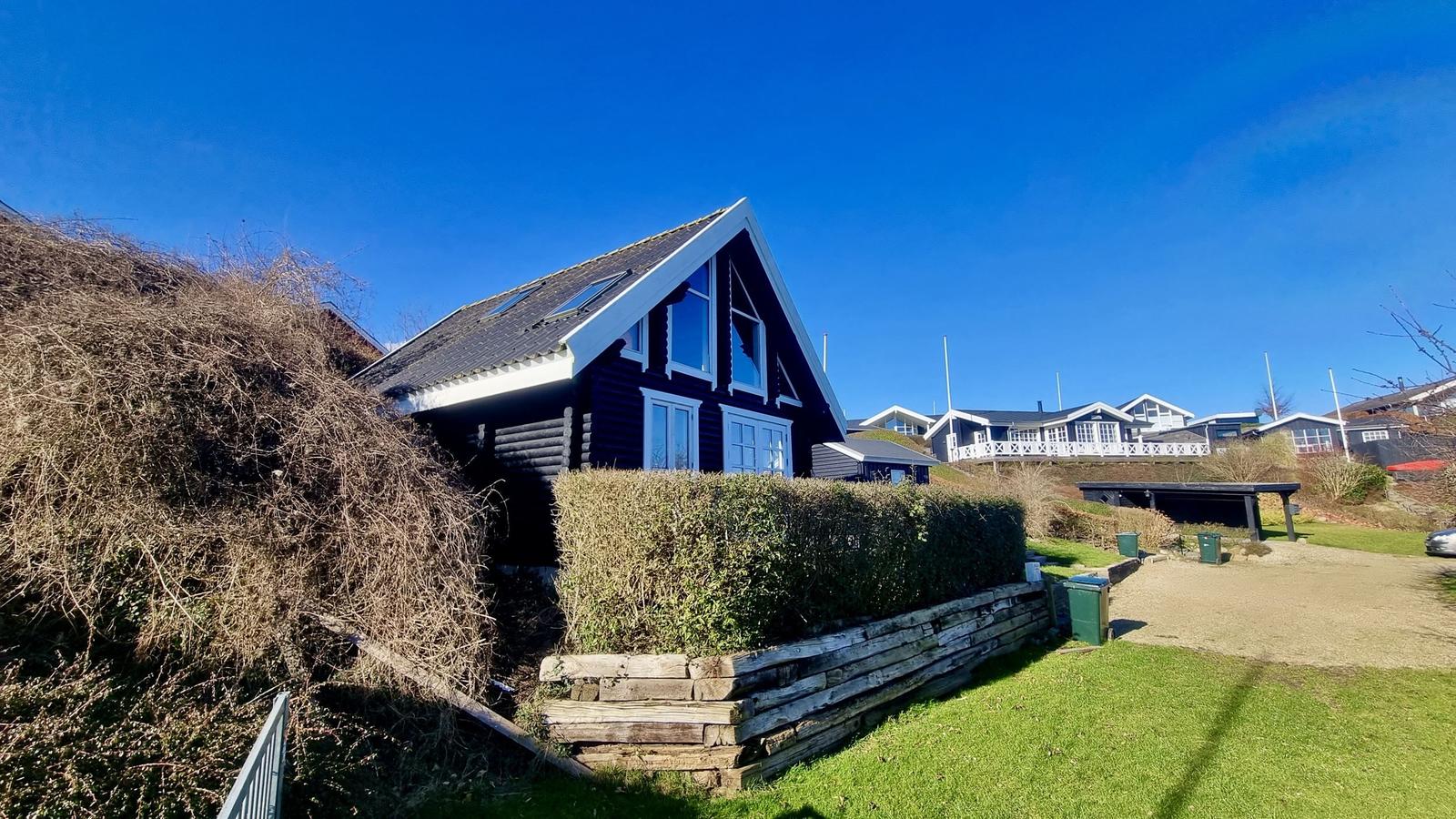 Schwarzes Holzhaus mit weißem Fensterrahmen und Dachgauben. Grünflächen und Hecke im Vordergrund.