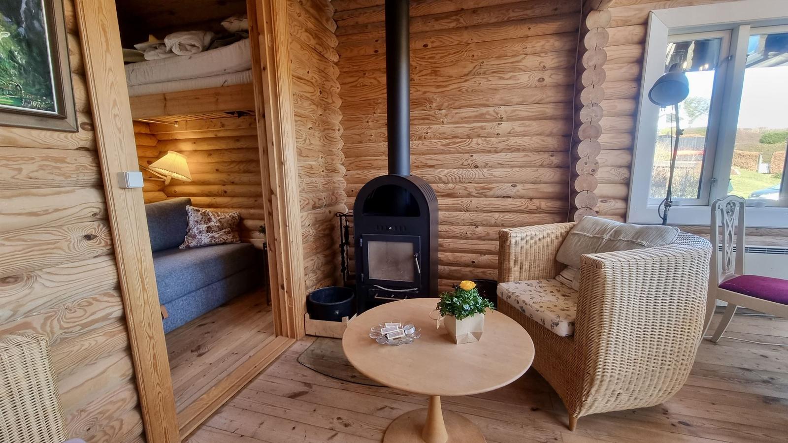 Wohnbereich mit Kamin, Sessel und Tisch. Im Hintergrund Schlafzimmer mit Sofa und Bett.