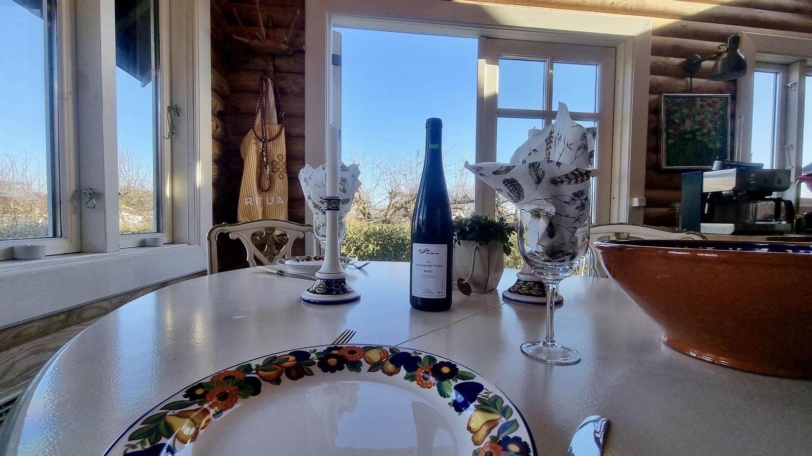 Tisch mit Teller, Weinflasche und Gläsern vor Fenster mit Blick auf Außenbereich.