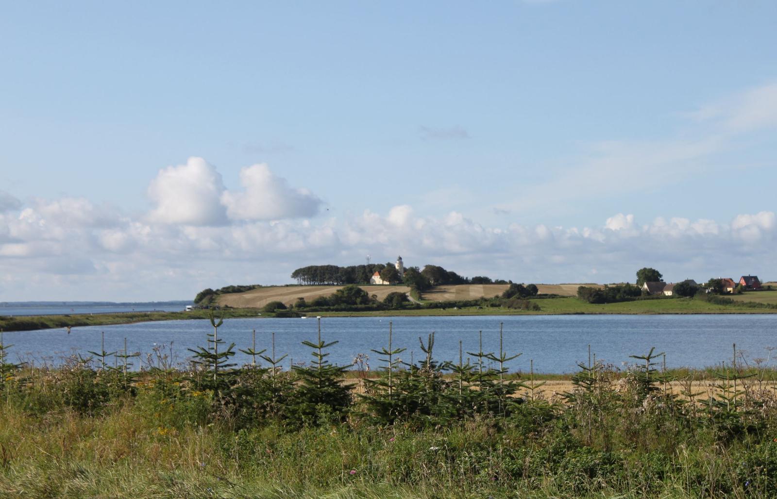 Blick auf einen See mit Insel, auf der ein Leuchtturm und Gebäude stehen.