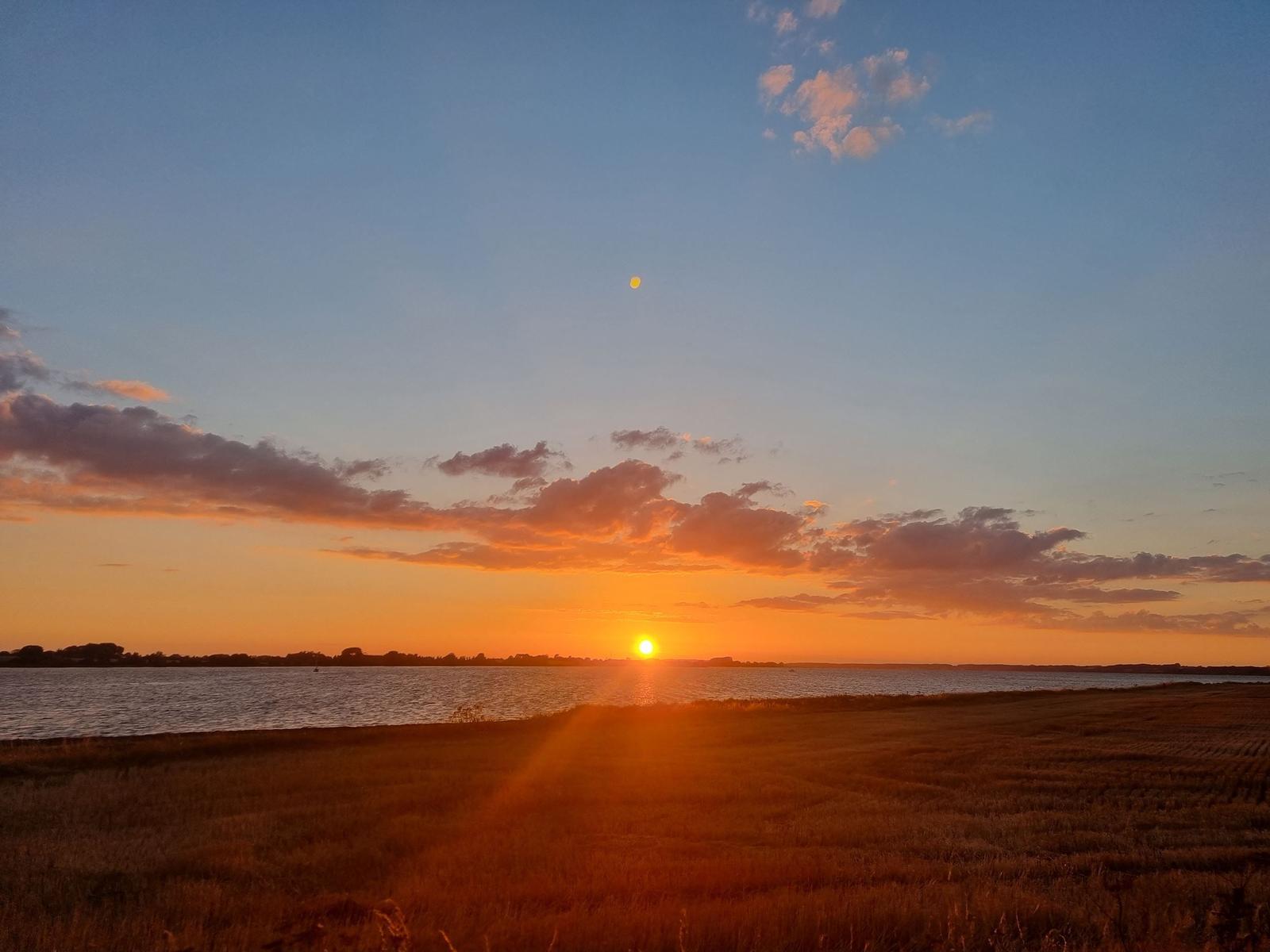Sonnenuntergang über See mit Feld im Vordergrund und Mond am Himmel.