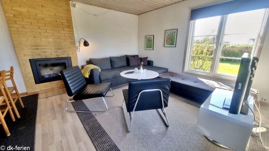 Wohnzimmer mit Sofa, Sitzgruppe und Kamin. Große Fenster mit Blick auf Grünfläche.