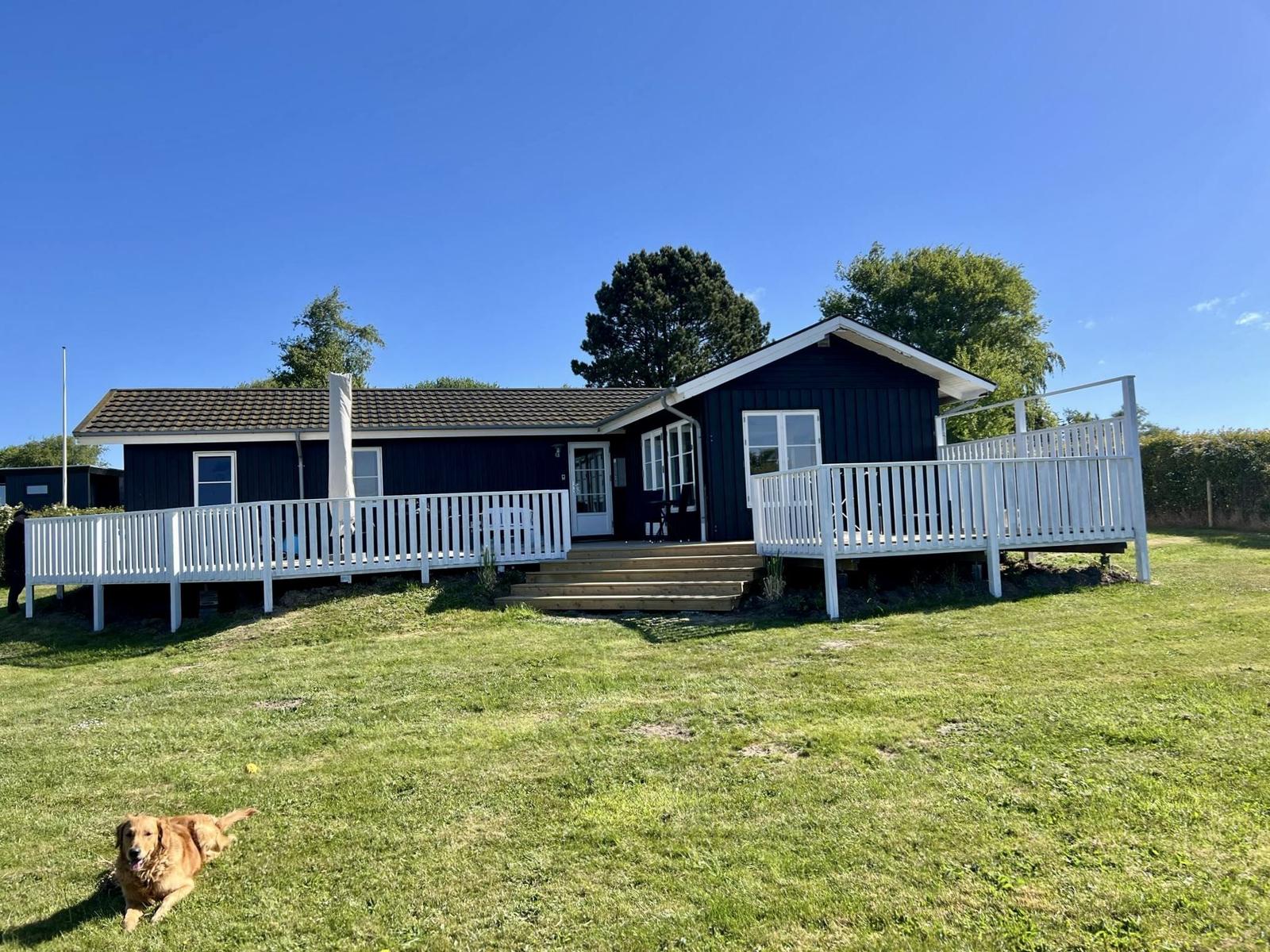 Schwarzes Ferienhaus mit weißer Veranda und Grünfläche. Ein Hund liegt im Vordergrund.