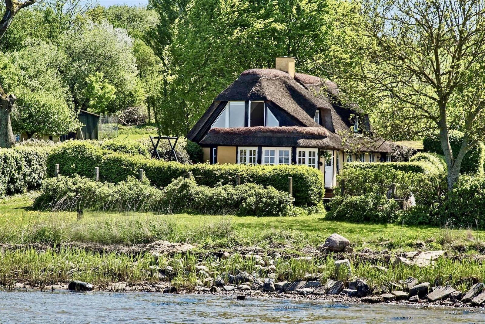 Hütte mit Strohdach am Wasser, umgeben von Grün und Bäumen.