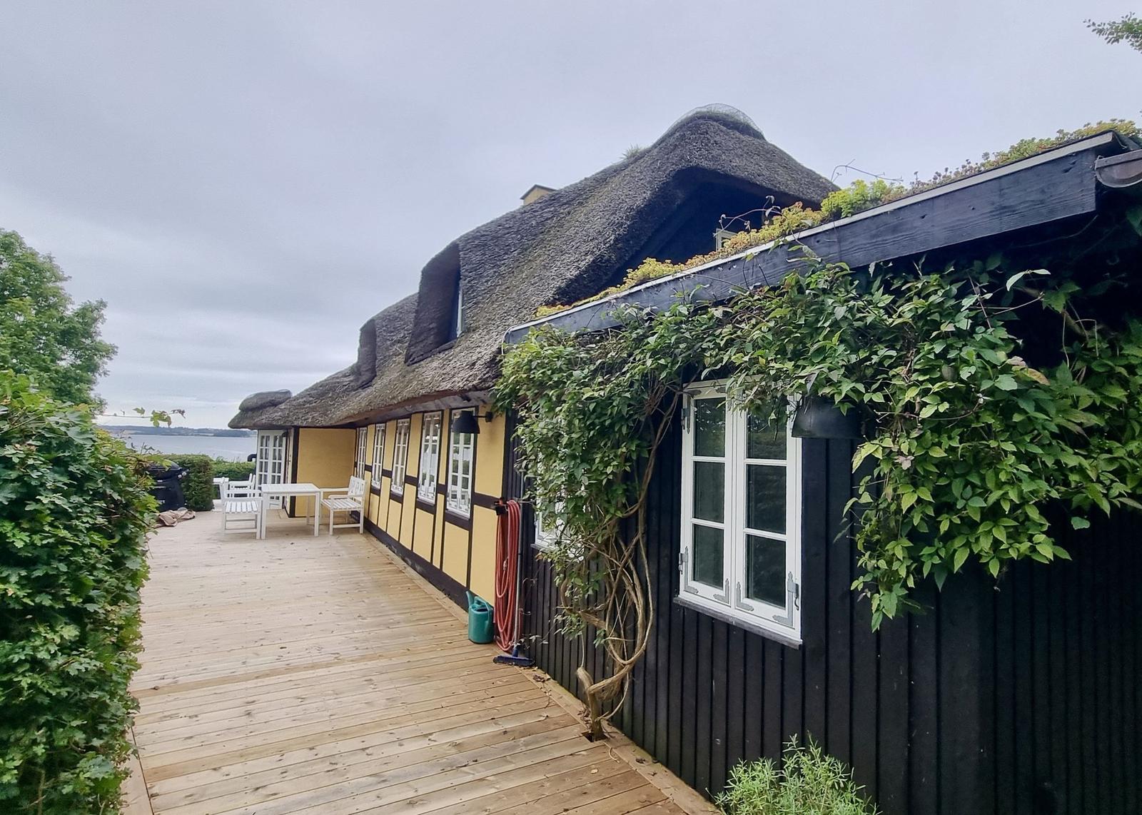 Haus mit Strohdach und Holzterrasse, umgeben von Grün und Wasser.