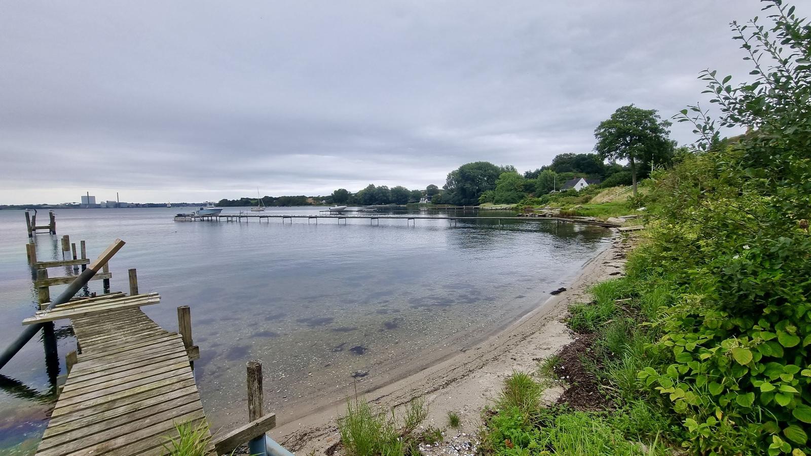 Holzsteg führt zum Wasser, Ufer mit Grünflächen und Bootsanlegestellen.