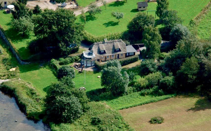 Dorfhaus mit Schieferdach, umgeben von Grünflächen und Bäumen. Flussufer im Vordergrund.