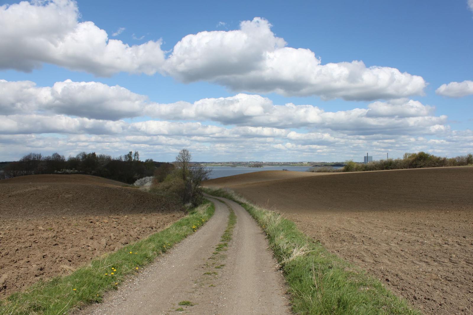 Dreckiger Weg durch Felder mit Blick auf See und Himmel mit Wolken.