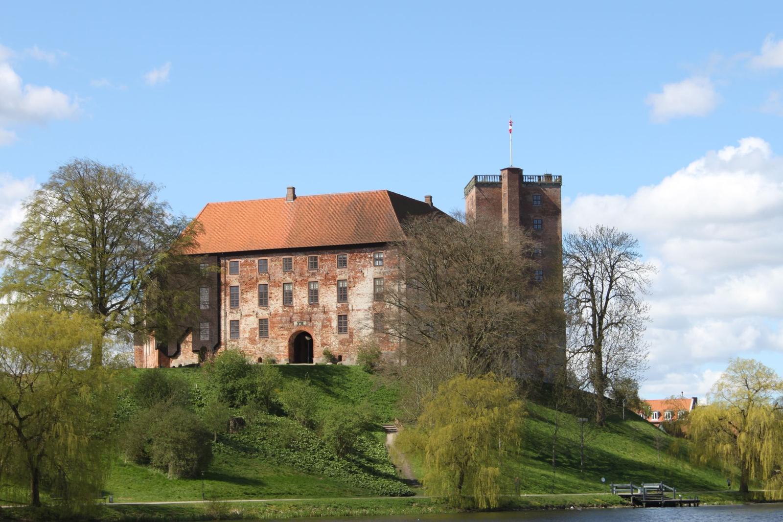 Schloss auf grüner Anhöhe mit Flagge, umgeben von Bäumen und Wasser.