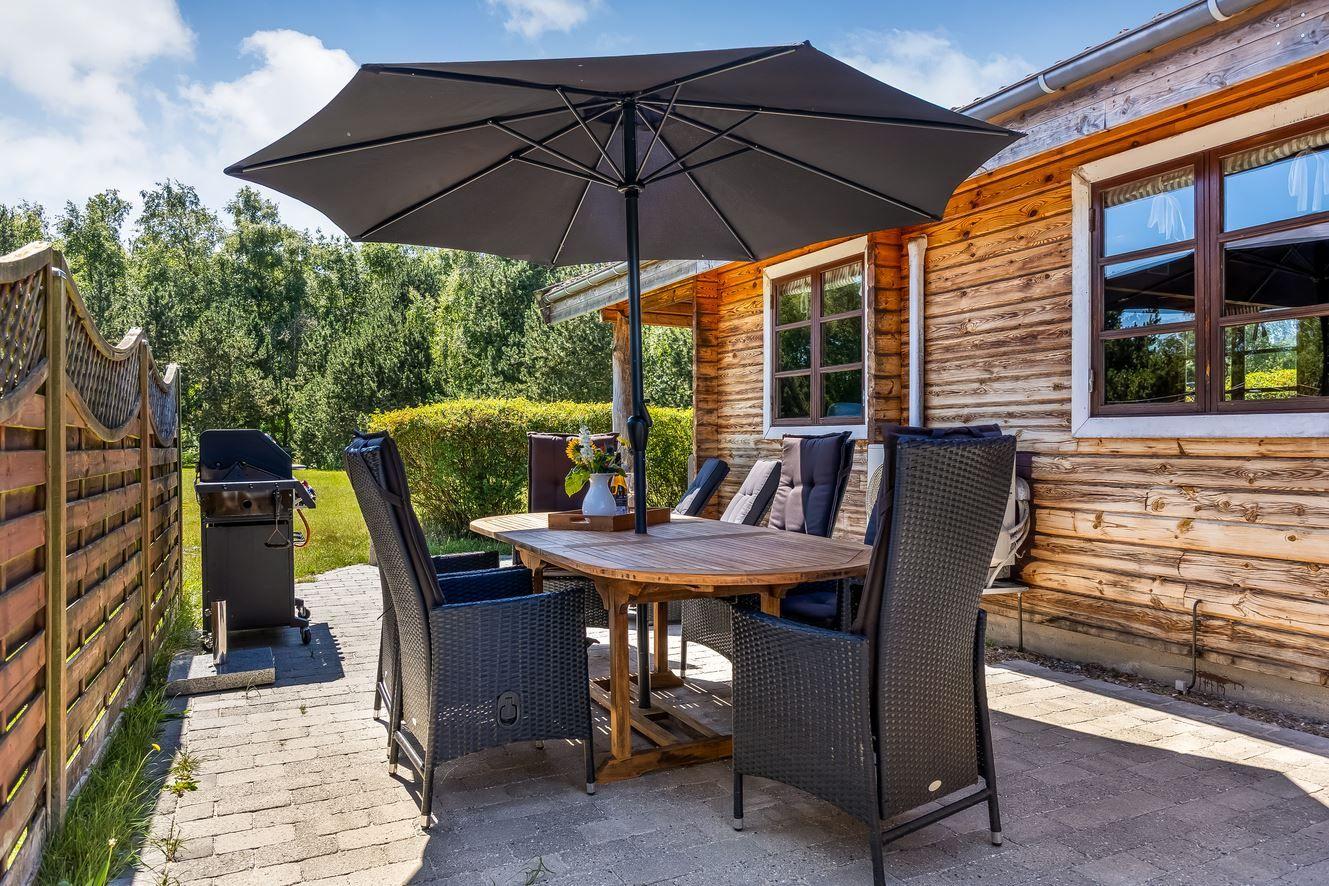 Terrasse mit Tisch, Stühlen und Sonnenschutz neben Holzhäusern.