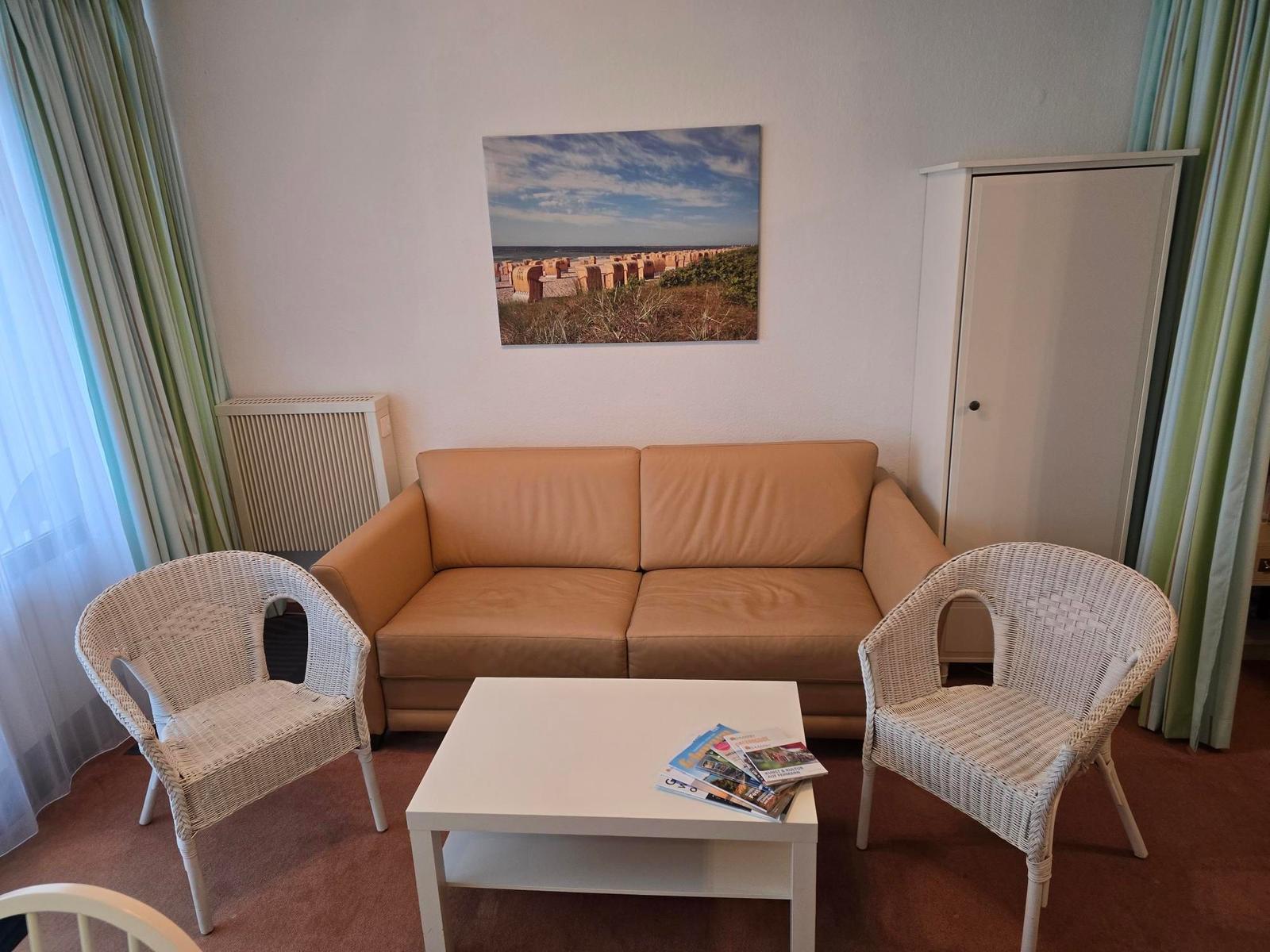 Ein Wohnzimmer mit Sofa, zwei Stühlen und Tisch. Wandbild und Gardinen sind sichtbar.