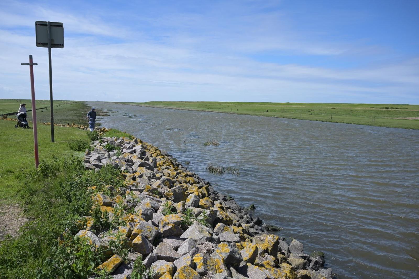 Wasserkanal mit Steinufer, Grasland und zwei Personen in der Ferne.