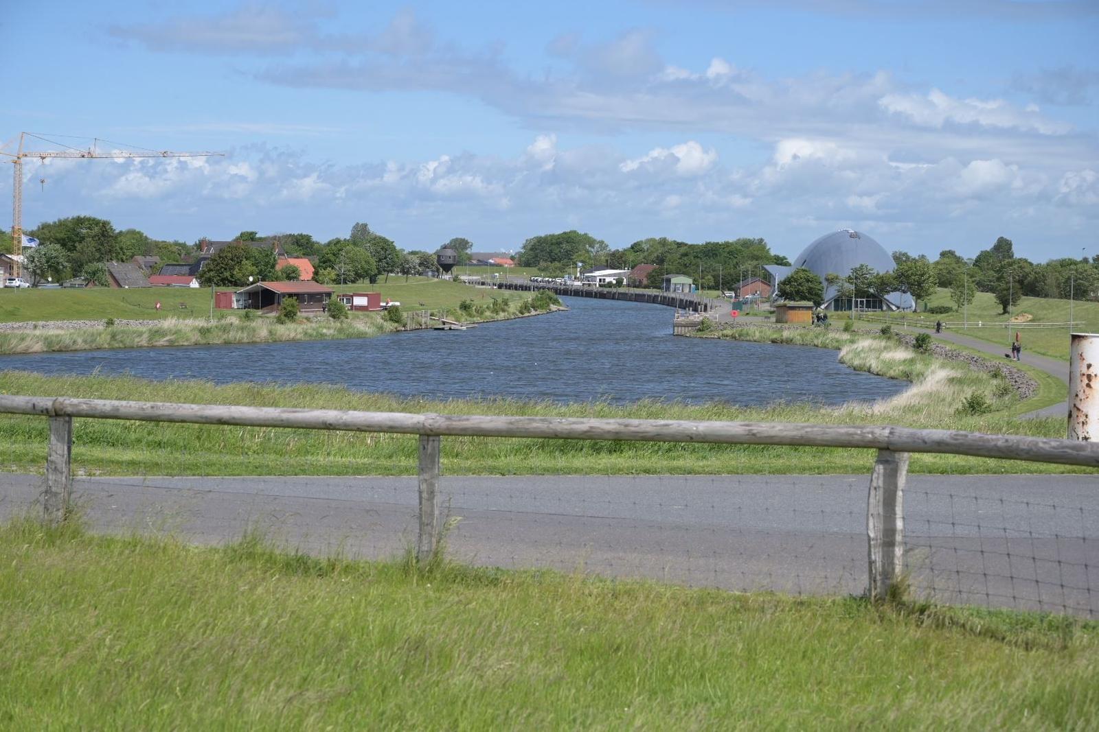 Flussufer mit Grünflächen, Weg und markanter Kuppelstruktur im Hintergrund.