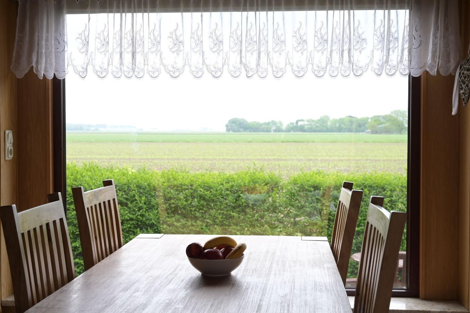 Esstisch mit Stühlen vor großem Fenster mit Blick auf Feld und Hecke.