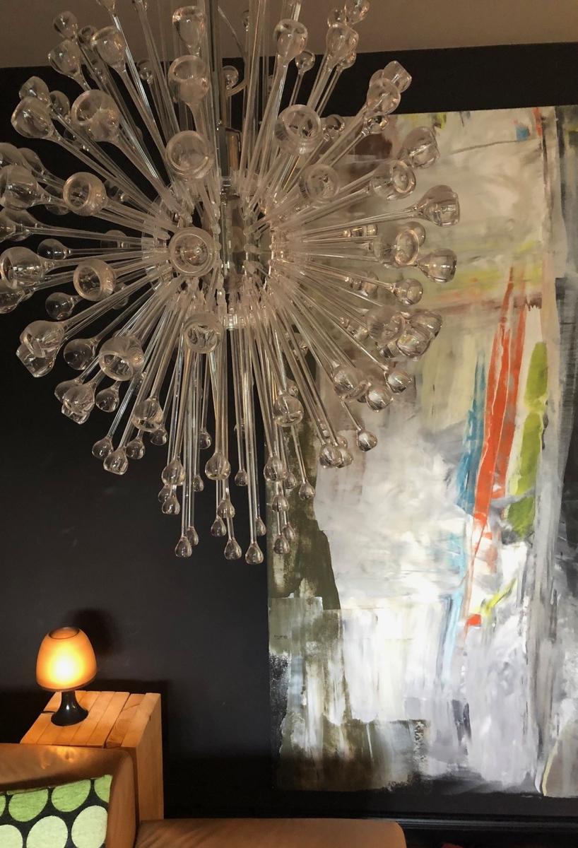 Ein Raum mit einer Glas-Kronleuchter, einer abstrakten Wandmalerei und einer kleinen Lampe auf einem Holztisch.