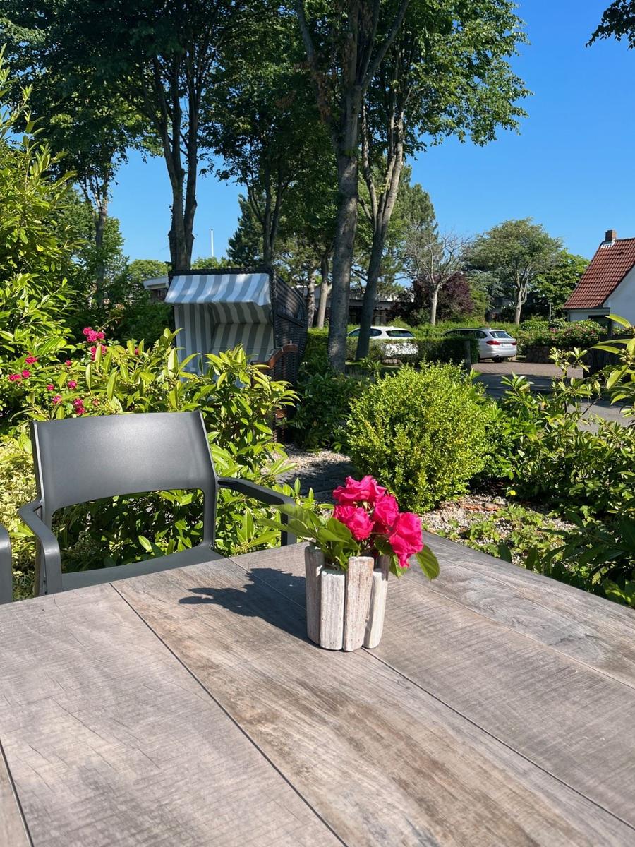 Holztisch mit Stuhl und Blumen im Garten. Im Hintergrund Bäume und ein Pavillon.