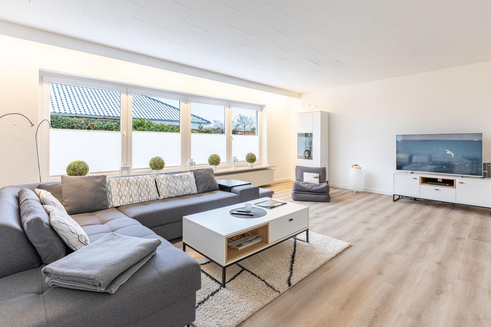 Wohnzimmer mit Sofa, Couchtisch und Fernseher. Große Fenster mit Blick auf Dach und Garten.