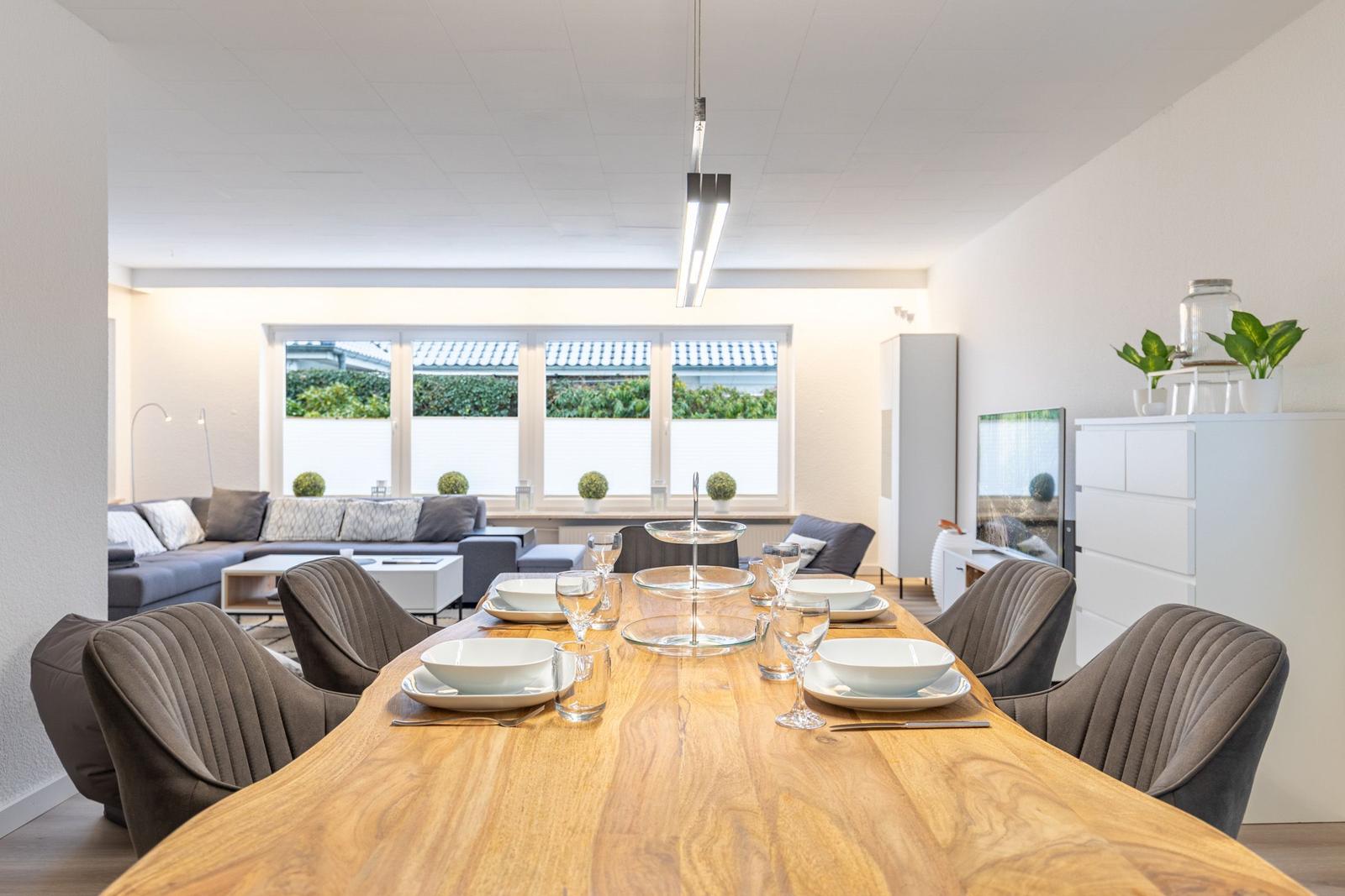 Essbereich mit Holztisch und Stühlen, dahinter Wohnzimmer mit Sofa und Fenster.